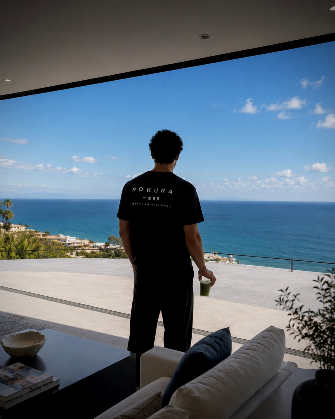 Man wearing a ROKURA t-shirt holding ceremonial matcha while overlooking the ocean from a modern minimalist luxury villa.